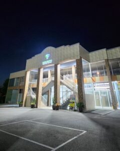 Exterior nighttime view of the two-story Medical Centre. The building features tan and stone-textured pillars, a central outdoor staircase leading to the second floor, and large glass storefronts on the ground level. A lighted sign with a green & blue logo is visible at the top center under a dark night sky.