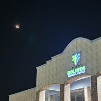 A nighttime architectural detail of the Holistic Medical Center showing the upper portion of the building and the illuminated "Holistic Medical Center" sign. The moon is visible in the upper left corner of the dark sky. The shot highlights the stone-textured pillars and the tan-patterned upper facade.