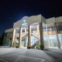 Exterior nighttime view of the two-story Medical Centre. The building features tan and stone-textured pillars, a central outdoor staircase leading to the second floor, and large glass storefronts on the ground level. A lighted sign with a green & blue logo is visible at the top center under a dark night sky.