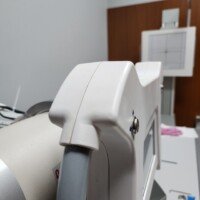 Gemini said Close-up of a medical X-ray machine head in a clinical room, showing the white plastic casing and handle, with a digital imaging detector panel and wooden wall paneling blurred in the background.