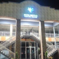 A close-up nighttime shot of the entrance to Holistic Medical Center, highlighting the symmetrical stone-textured pillars and the illuminated brand logo above the second-story balcony. Potted plants flank the ground-floor glass doors, and the white-railed staircases are visible on either side.