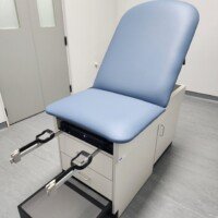 A close-up of a blue and white adjustable medical examination table with pull-out footrests and storage drawers in a clinical room.