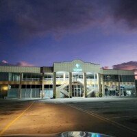 A view of the Holistic Medical Center at dusk from inside a vehicle. The purple and blue sunset sky is filled with clouds above the two-story medical facility. The building's logo is illuminated, and the empty parking lot is visible in the foreground.