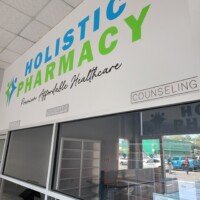 A bright interior view of the Holistic Pharmacy. Above the service counter, a large wall graphic reads "HOLISTIC PHARMACY" in blue and green letters with the slogan "Premium Affordable Healthcare" underneath. Signs for "Dispensary" and "Counseling" are visible above glass service windows.