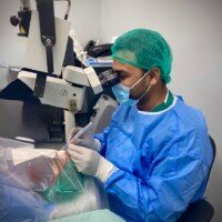 A surgeon in blue scrubs, a mask, and a hairnet performs a precise procedure on a patient's eye using a large medical microscope.