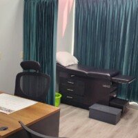 A combined medical office and exam room. On the left is a wooden desk with a computer and chair; on the right, behind a teal privacy curtain, is a black padded examination table with integrated storage drawers.