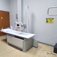A radiology room featuring a padded medical imaging table and a ceiling-mounted X-ray tube. A yellow "Caution" sign on the wall warns that pregnant women should not enter.