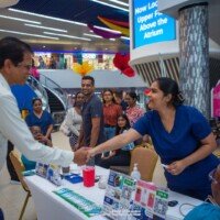 A healthcare worker in blue scrubs shakes hands with a man at a public health screening booth in a brightly lit atrium.