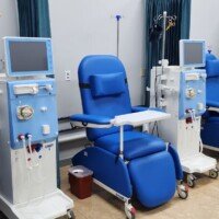 A row of several blue clinical dialysis chairs, each paired with a white-and-blue medical unit featuring a monitor and IV pole. The treatment stations are separated by dark teal privacy curtains.