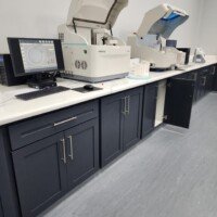 A high-tech medical laboratory featuring automated diagnostic machines and computer monitors on a clean white countertop with dark cabinetry.
