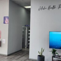 A wide view of a medical center's modern reception area. A large wall features a clock with word-based numbers and the text "Holistic Health: The Whole You Matters." The space includes a flat-screen TV displaying a landscape, potted plants, a hand sanitizer station, and parked wheelchairs.