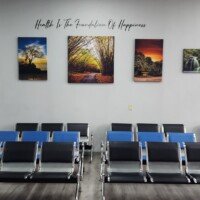 A medical facility waiting room with rows of blue and black metal chairs. The wall behind the seating features several nature-themed canvas prints and a quote that reads "Health Is The Foundation Of Happiness."