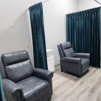 Two grey leather recliner chairs are positioned in semi-private bays divided by dark green privacy curtains. The room features grey wood-style flooring and neutral grey walls.