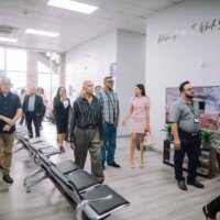Patients and visitors walking through a bright, spacious medical center lobby featuring blue seating and a wall sign that reads "Holistic Health... The Whole You Matters."
