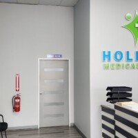 modern medical reception area featuring a large "Holistic Medical Center" logo on a grey wall. The space includes a black-and-white striped desk with informational brochures and a red fire extinguisher mounted on the adjacent wall.